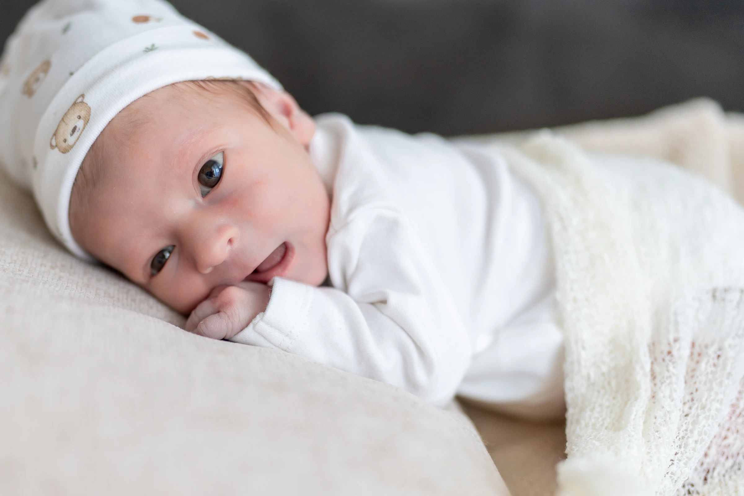 Waches Baby mit weisser Mütze liegt auf einer cremefarbenen Decke und schaut in die Kamera